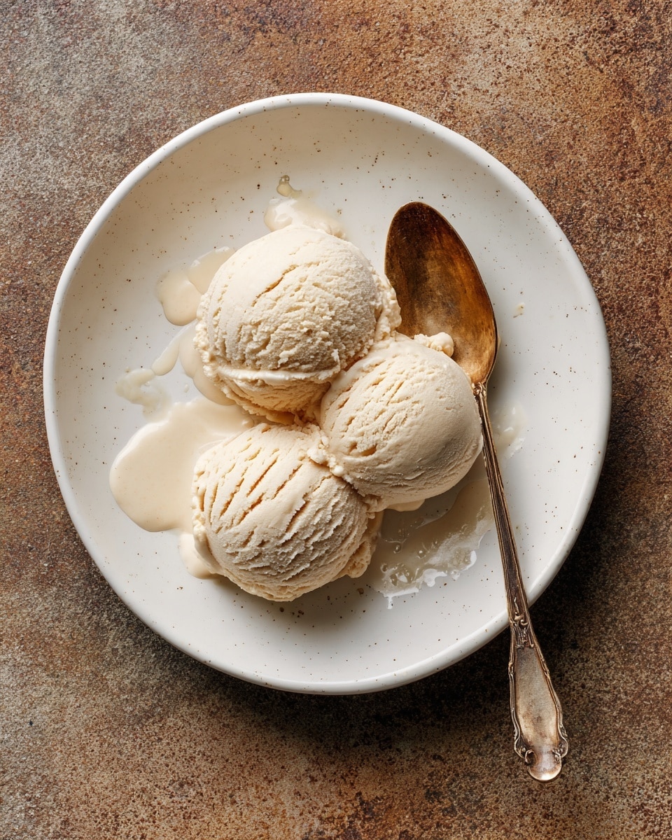 A white plate holds three light beige scoops of ice cream, each textured with soft cracks and smooth curves. The scoops are closely grouped near the center-left of the plate, with small drips of melted ice cream around them adding a shiny, wet detail. A tarnished golden spoon rests on the right side of the plate, its surface showing a matte, aged finish. The plate sits on a white marbled surface that adds a soft, elegant texture to the background. photo taken with an iphone --ar 4:5 --v 7