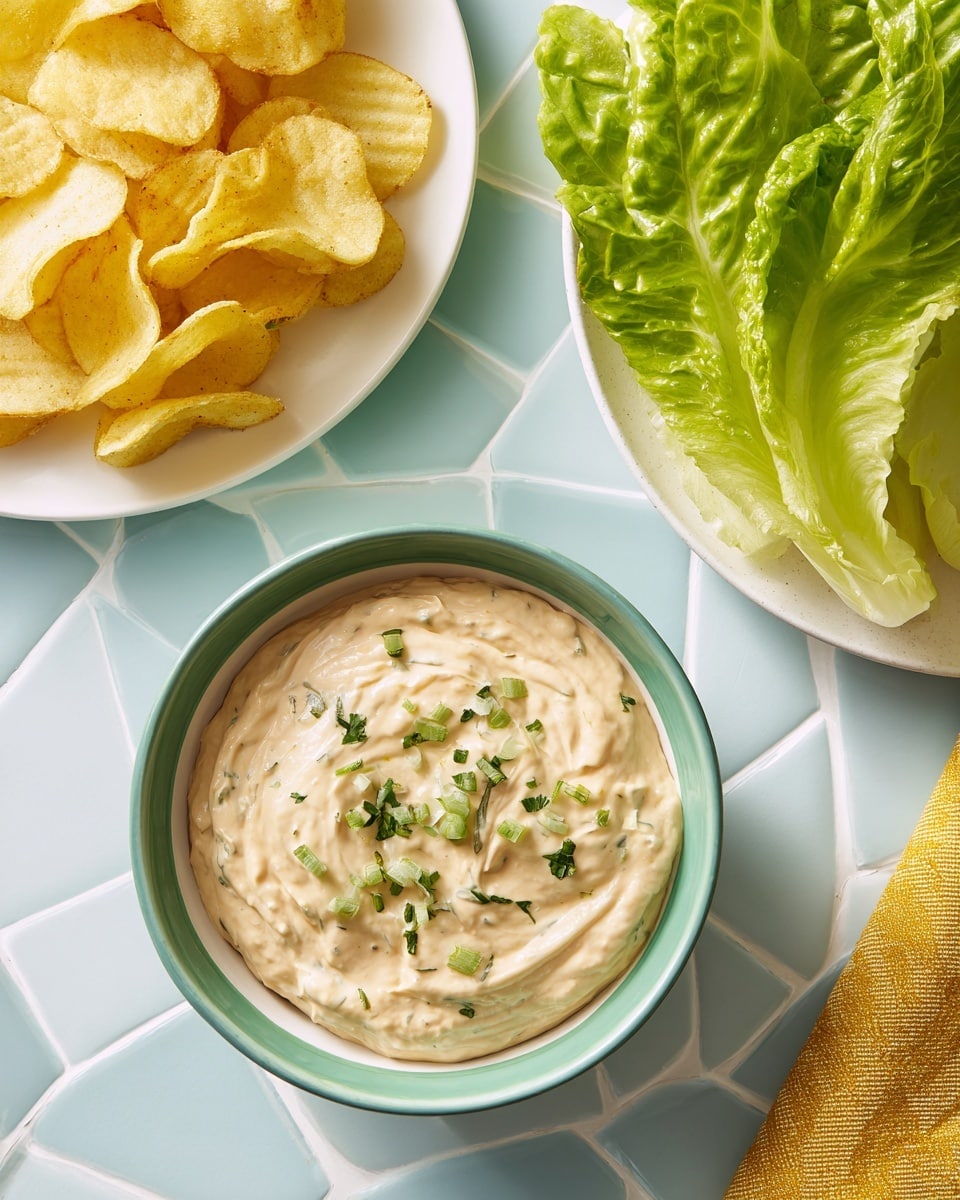 A white bowl is filled with smooth, light beige dip topped with small green herb pieces, placed on a white marbled surface. A woman's hand holds a ridged potato chip partially dipped in the creamy dip. In the background, there is a white bowl filled with more ridged potato chips on the left and a white plate with fresh green lettuce on the right, both on the same white marbled surface. photo taken with an iphone --ar 4:5 --v 7