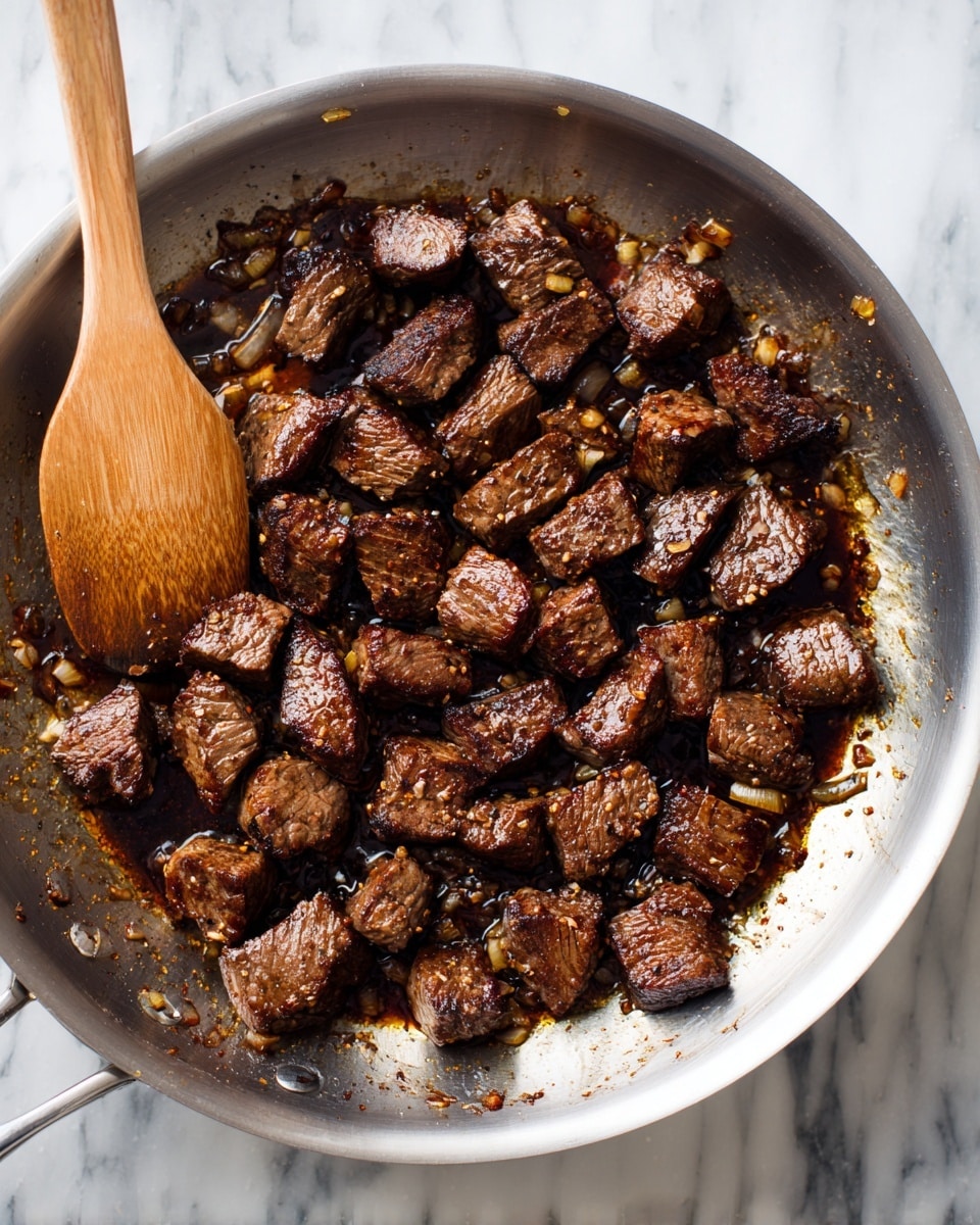 The image shows a silver pan filled with many small pieces of cooked meat, dark brown in color with a seared texture. The meat pieces are irregular in shape and spread evenly across the pan, which has bits of caramelized onions and browned juices scattered around them, creating a shiny, rich sauce. A wooden spatula is resting inside the pan on the left side, slightly lifting some pieces of meat. The background surface has a white marbled texture. photo taken with an iphone --ar 4:5 --v 7