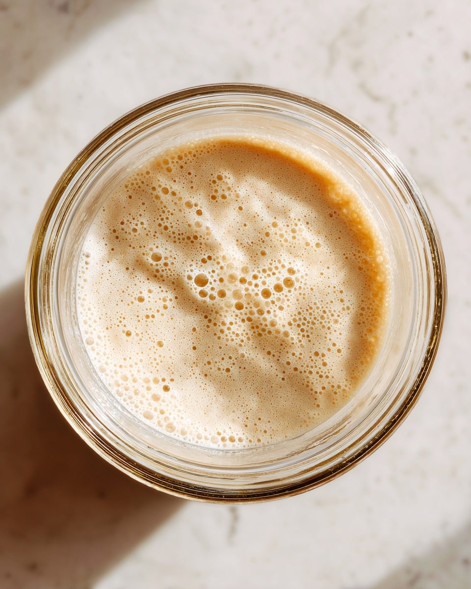 A close-up top view of a blender jar filled with a creamy light beige mixture that looks smooth and frothy. The inside of the jar is clear with soft light reflections, showing the texture of the blended content clearly. The blender jar sits on a surface with a white marbled texture, and the image has a bright, clean feel. photo taken with an iphone --ar 4:5 --v 7